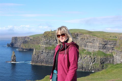 Blonde woman in fuchsia jacket and sunglasses smiles before cliffs overlooking the sea