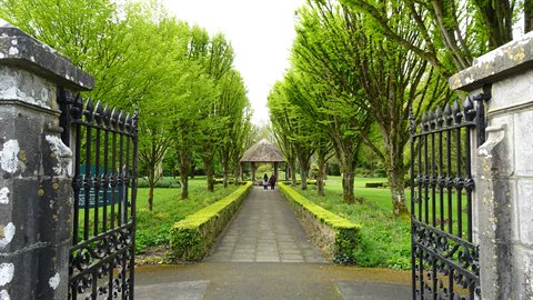 Wrought iron gate between stone pillars, tree-lined path toward a thatched pavilion