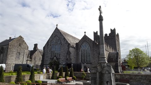 Medieval grey stone church with tower, Celtic cross in foreground, cloudy sky, tourists walking