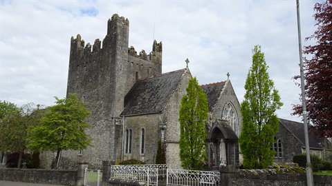 Medieval stone church with crenellated tower, white gates, green trees, cloudy sky