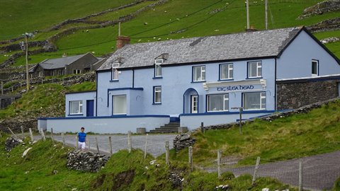 Person walking toward a light blue pub on a green hillside, soft light