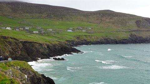 Tourist photographs the rocky coast with scattered houses on green slopes under cloudy sky