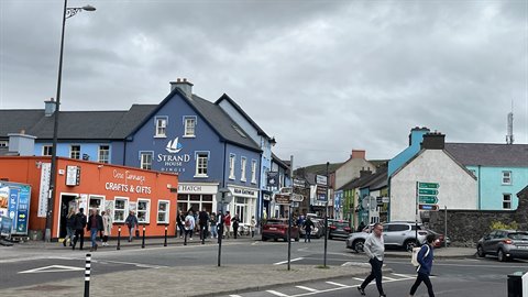 Pedestrians cross a town street lined with colorful shops under cloudy sky