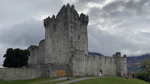 Ancient grey stone castle under cloudy sky, visitors walking on green lawn