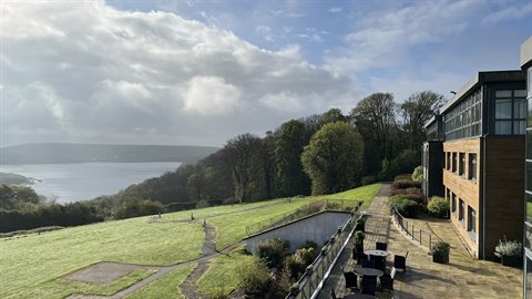 Tables on terrace before modern hotel, bay and wooded hills under clouds