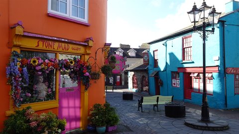 Small square with orange and blue painted shops, flower displays and green bench in daylight