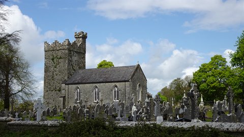 Stone church with medieval tower among Celtic gravestones and green trees, cloudy sky