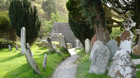 Gravel path between leaning old gravestones and stone chapel, dappled sunlight