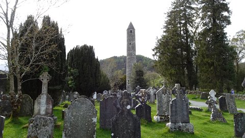Stone round tower among Celtic gravestones and tall trees in ancient monastic graveyard