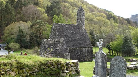 Stone chapel with bell tower, Celtic cross and gravestones, green woods, daylight