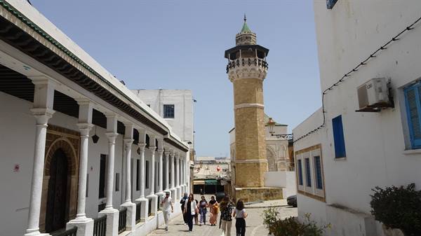 Tourists walk along an arcaded white alley toward a stone minaret