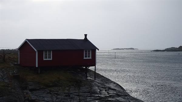 Red wooden cabin on rocky shore by the calm sea, under grey sky