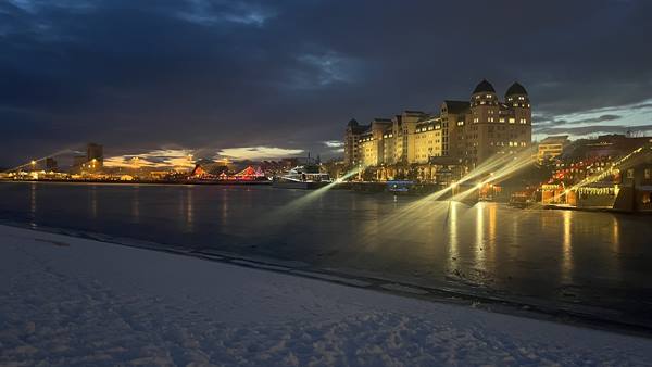 Snowy shore, illuminated building reflecting lights on water at dusk