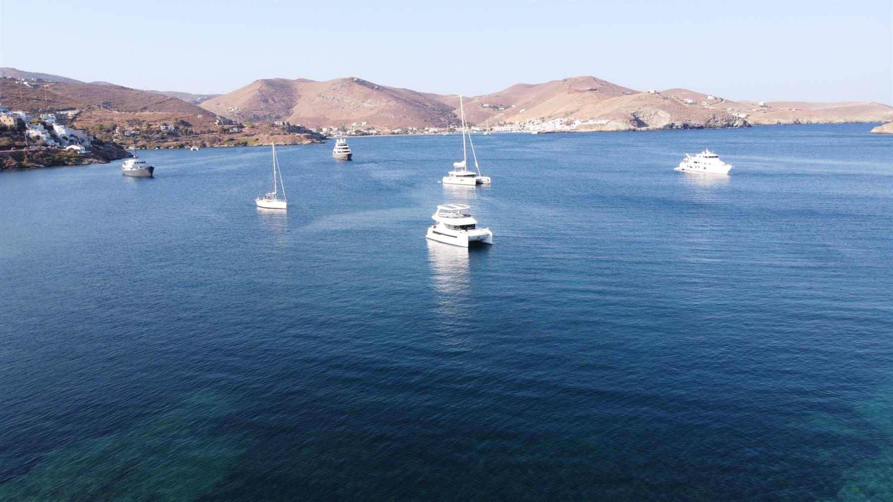 Barche a vela e yacht ancorati in baia turchese, colline aride sullo sfondo