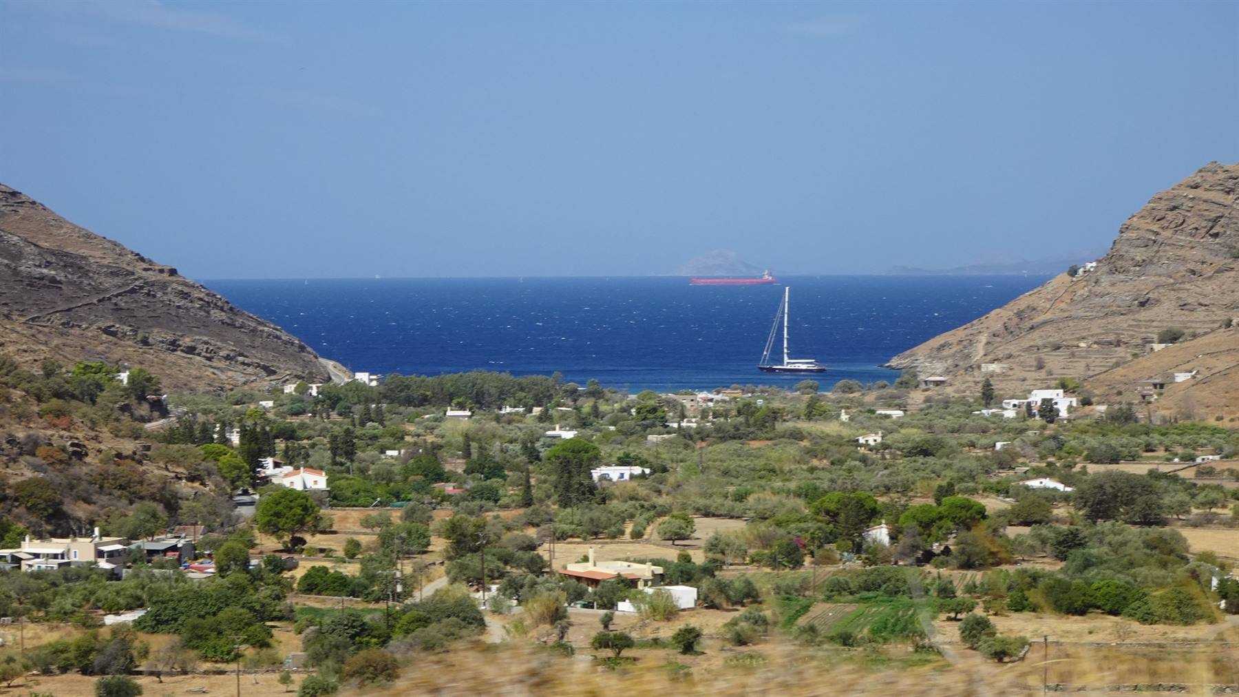 Valle verde tra colline aride, case bianche e barca a vela sul mare blu