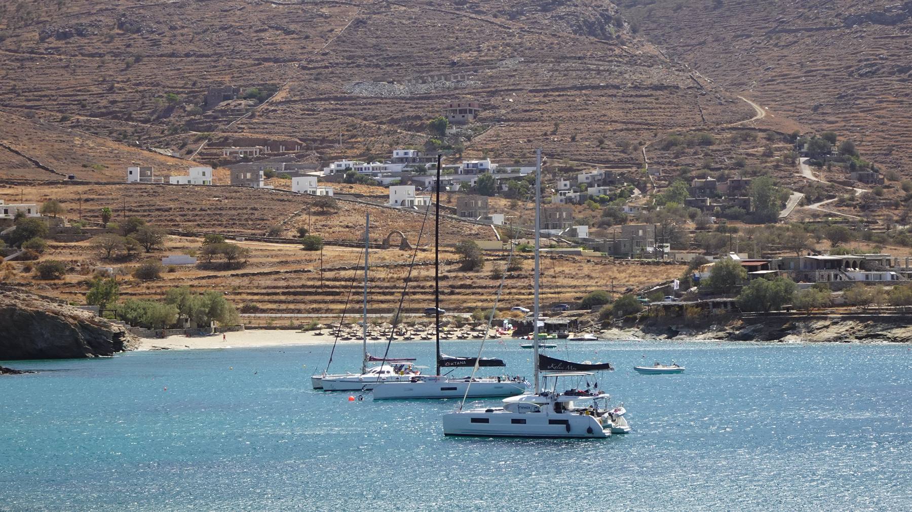 Catamarani e barche a vela ancorati in baia turchese sotto colline aride con case bianche