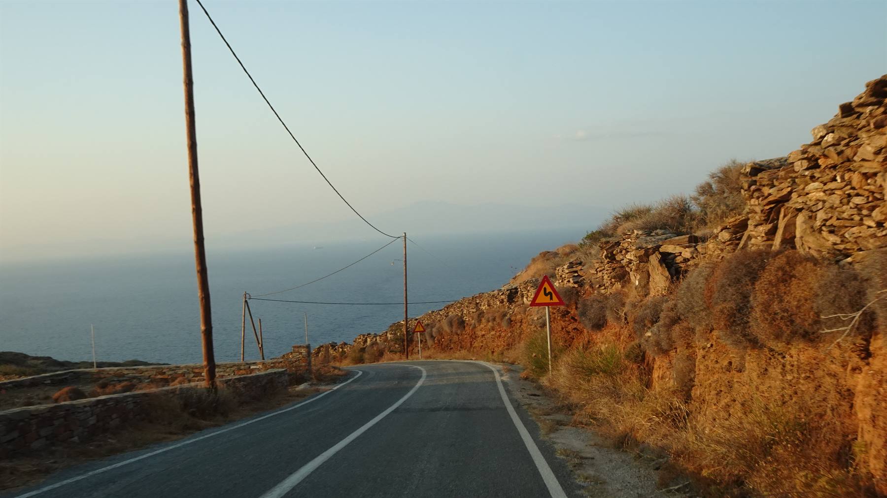 Strada asfaltata verso il mare tra muretti in pietra e cespugli aridi, luce dorata
