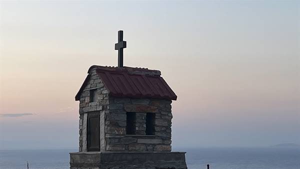 Small stone chapel with red roof and cross, by the sea at dawn