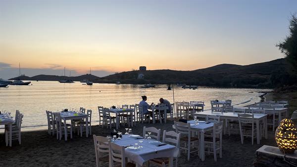 Set tables on the beach at dusk, sailboats anchored in the bay