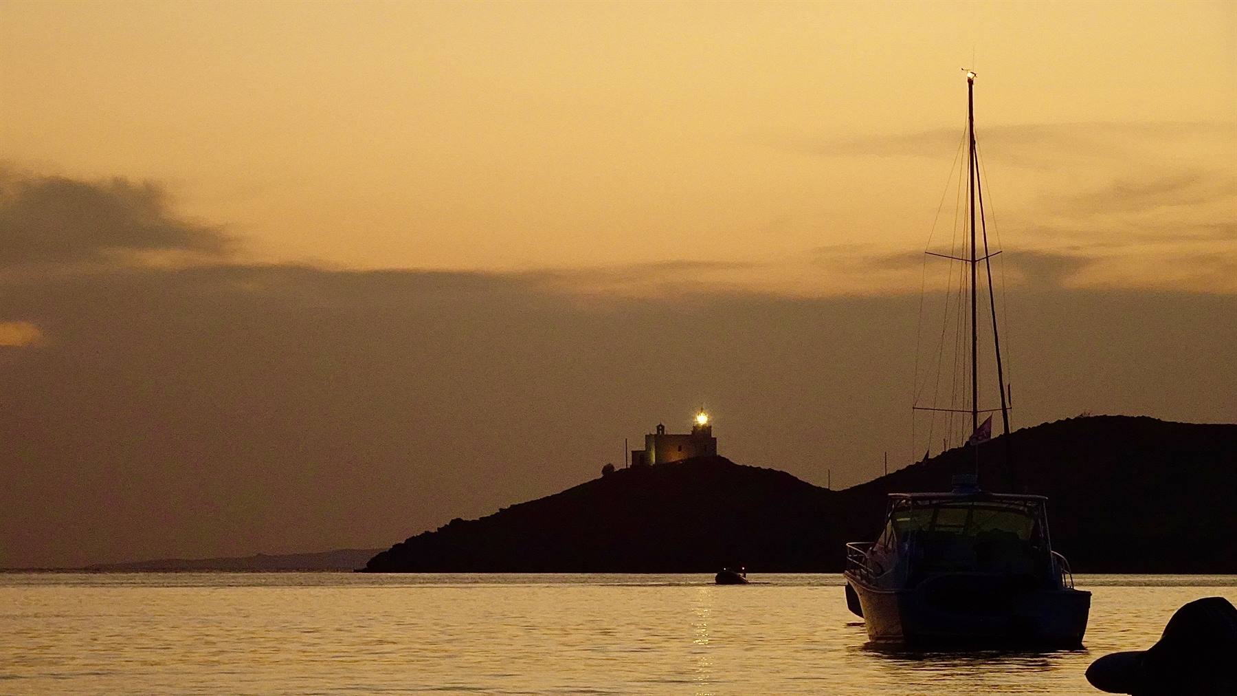 Barca a vela ancorata al largo, faro illuminato su collina scura contro cielo arancio