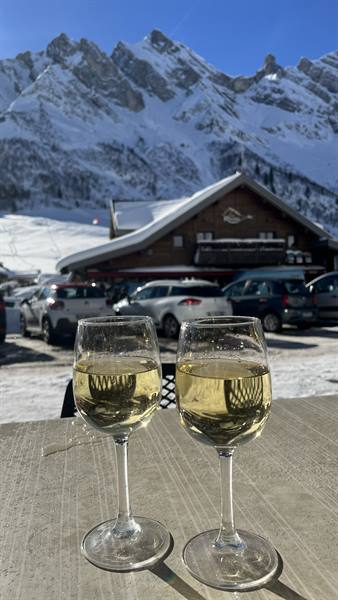 Two glasses of white wine on stone table, snowy chalet and peaks at Col des Aravis