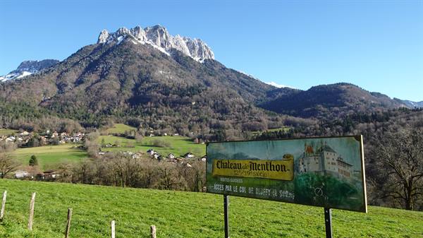 Château de Menthon billboard on green meadow, village and sunny rocky mountains