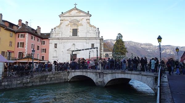 Crowd on stone bridge before Annecy church, Christmas lights, mountains behind