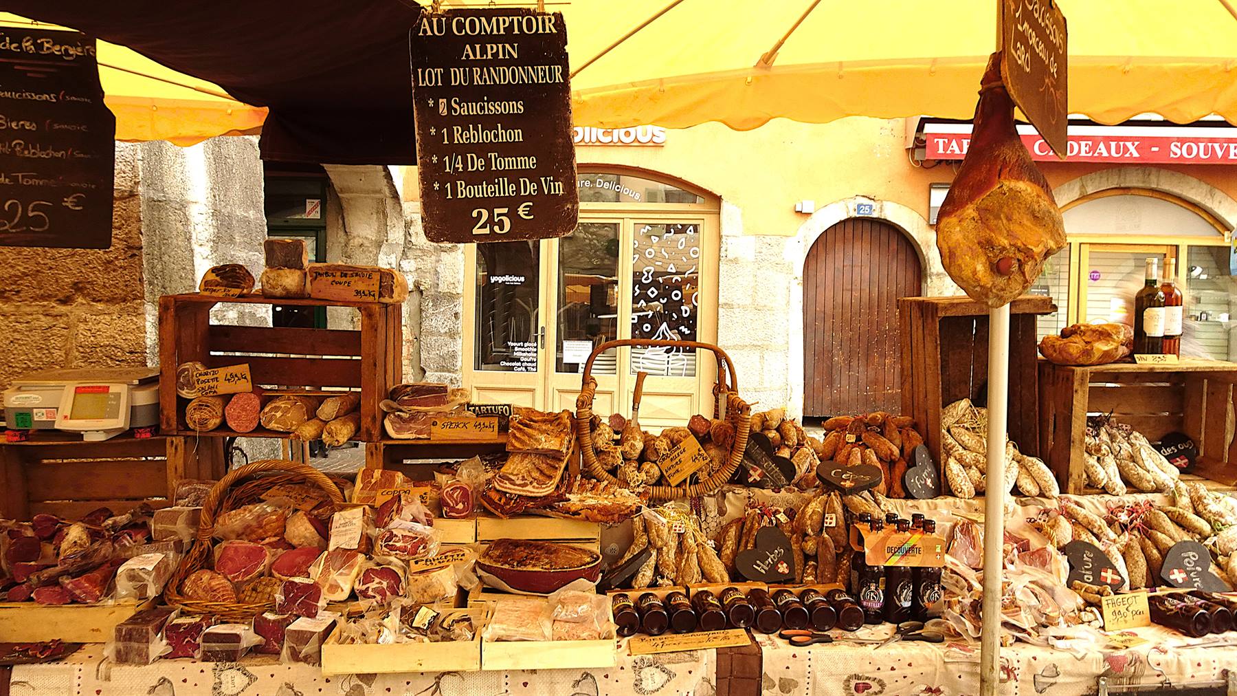 Banco di salumi e formaggi sotto ombrellone giallo, insegna Au Comptoir Alpin