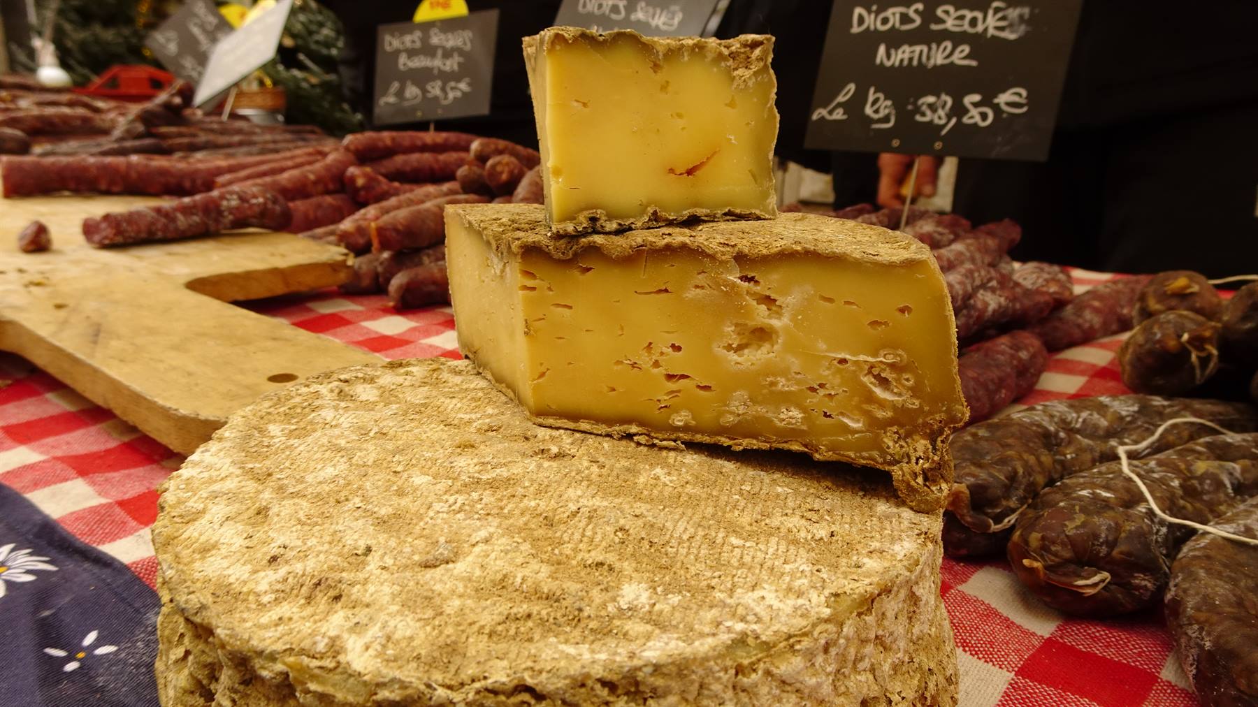 Formaggi e salumi su banco di mercato con tovaglia a quadri rossa, lavagna coi prezzi