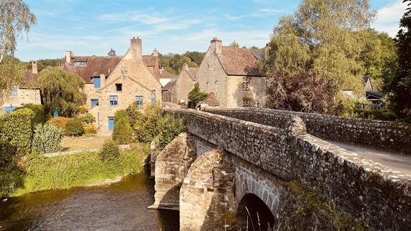 Medieval stone bridge over the river in Saint Céneri-le-Gérei, sunlit houses and trees