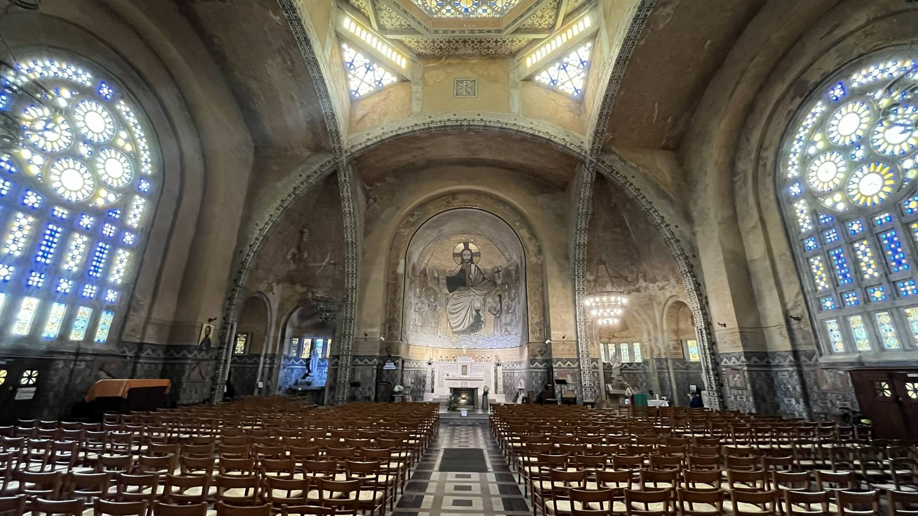 Interno di chiesa con sedie in legno, vetrate policrome e mosaico sopra l'altare