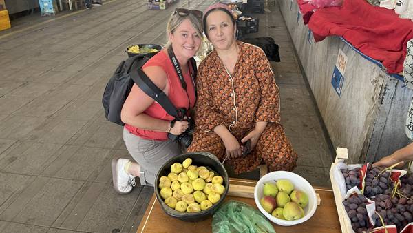 Due turiste con macchina fotografica accanto a una venditrice di frutta al bazar Chorsu di Tashkent