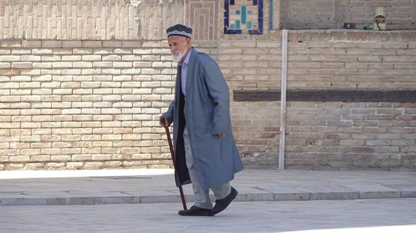 Anziano con camminello e veste grigia cammina lungo il muro di mattoni a Khiva