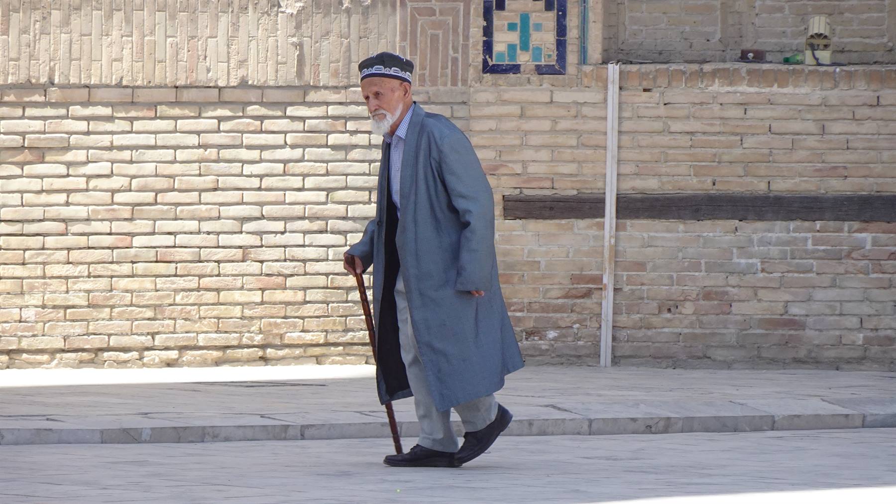 Anziano con camminello e veste grigia cammina lungo il muro di mattoni a Khiva