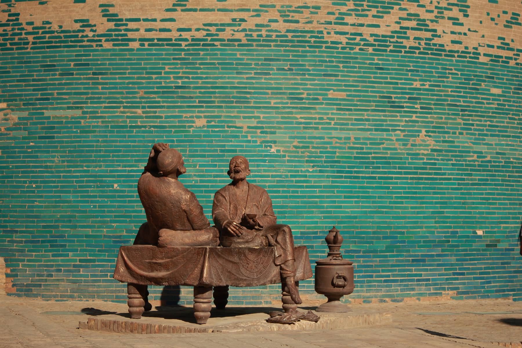 Statue in bronzo su una panca davanti alle maioliche turchesi del minareto Kalta Minor, Khiva