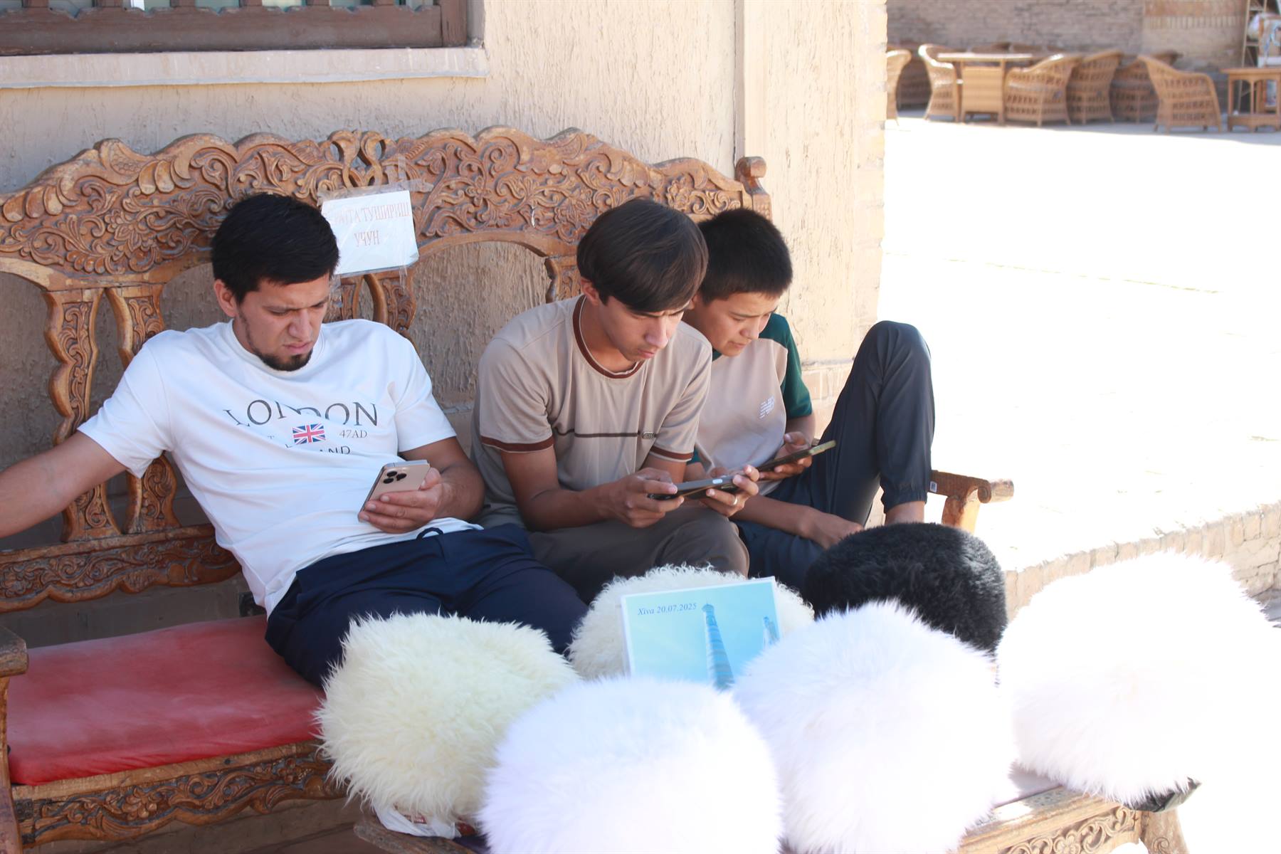 Ragazzi al telefono su panca intagliata in legno tra cappelli di pelliccia, Khiva