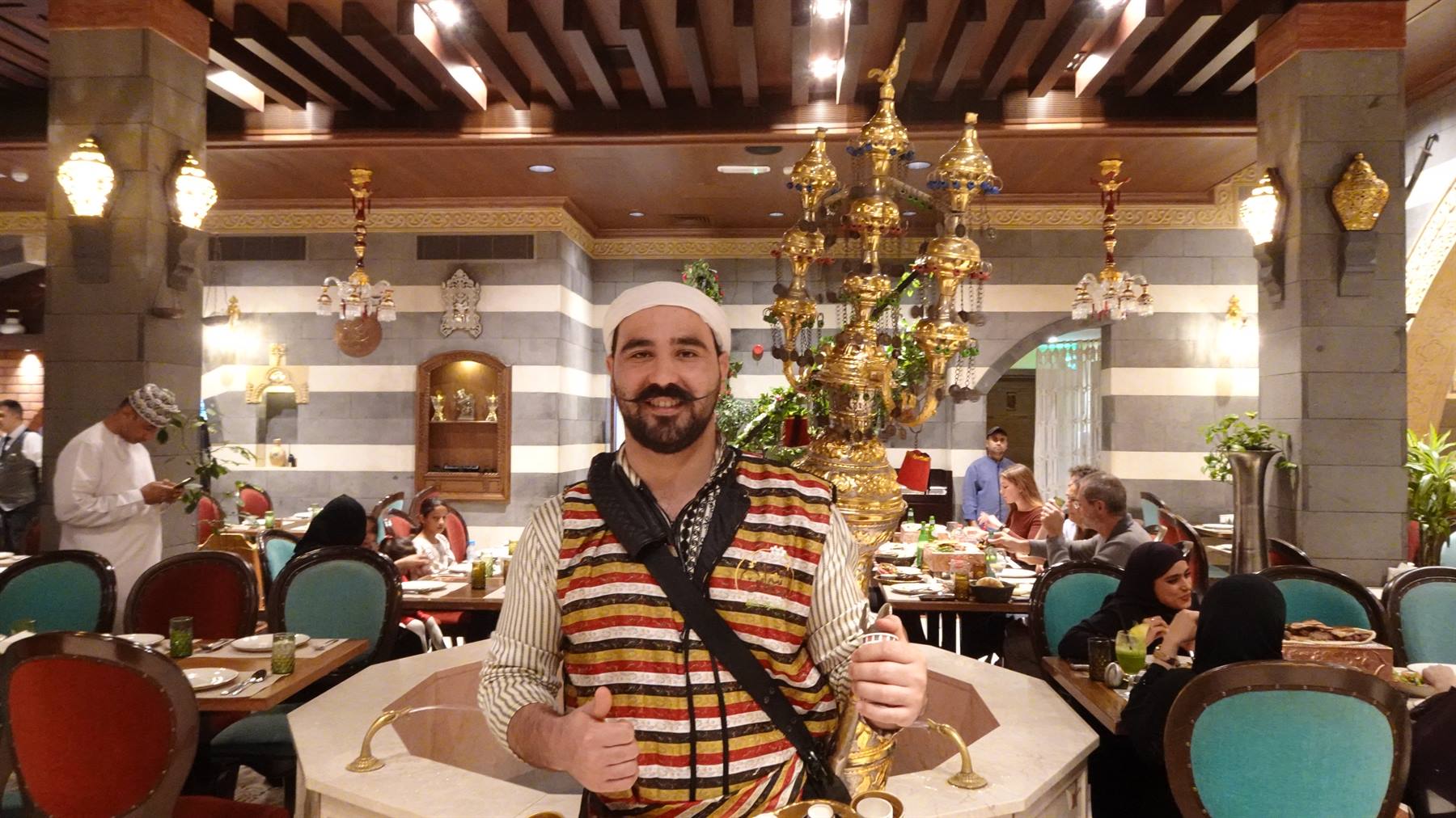 Cameriere in costume tradizionale sorride accanto a un lampadaio d'ottone nel ristorante