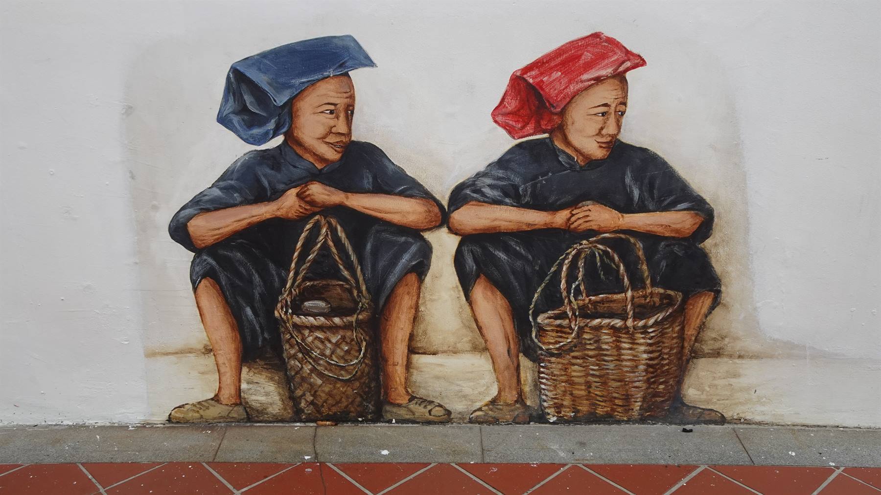 Murale di due donne sedute con cesti di vimini, cappelli blu e rosso, su muro bianco