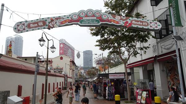 Chinatown Street Market sign above busy street, shoppers at stalls, skyscrapers behind