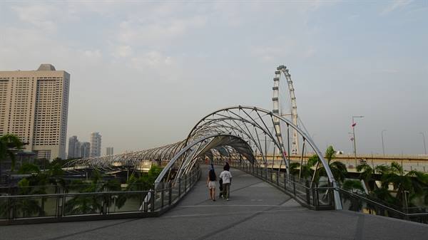 People walk across the steel Helix Bridge with the Singapore Flyer wheel behind