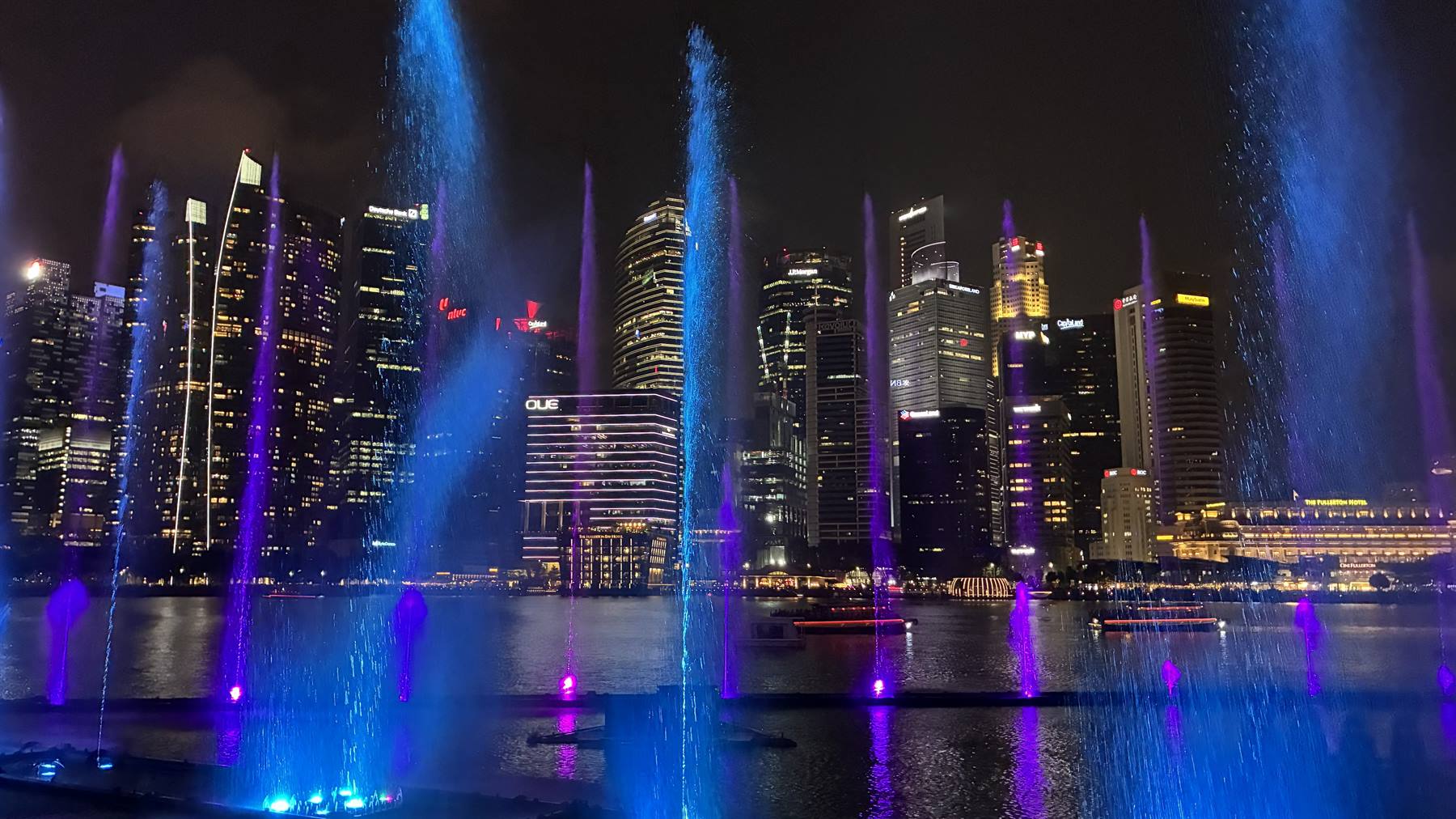 Getti d'acqua blu e viola illuminati su Marina Bay tra grattacieli notturni