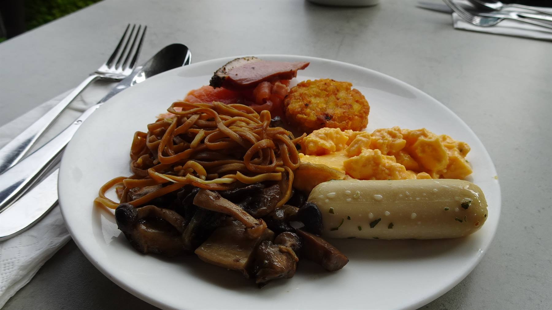 Piatto bianco con uova strapazzate, noodle, funghi e salsiccia, posate sul tavolo