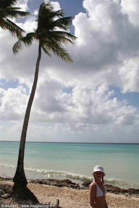 Tourist in white hat and bikini on beach under a leaning palm, turquoise sea