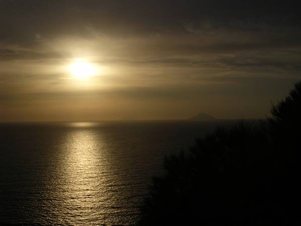 Low sun reflecting on the sea, Stromboli silhouette on the horizon, dark pines