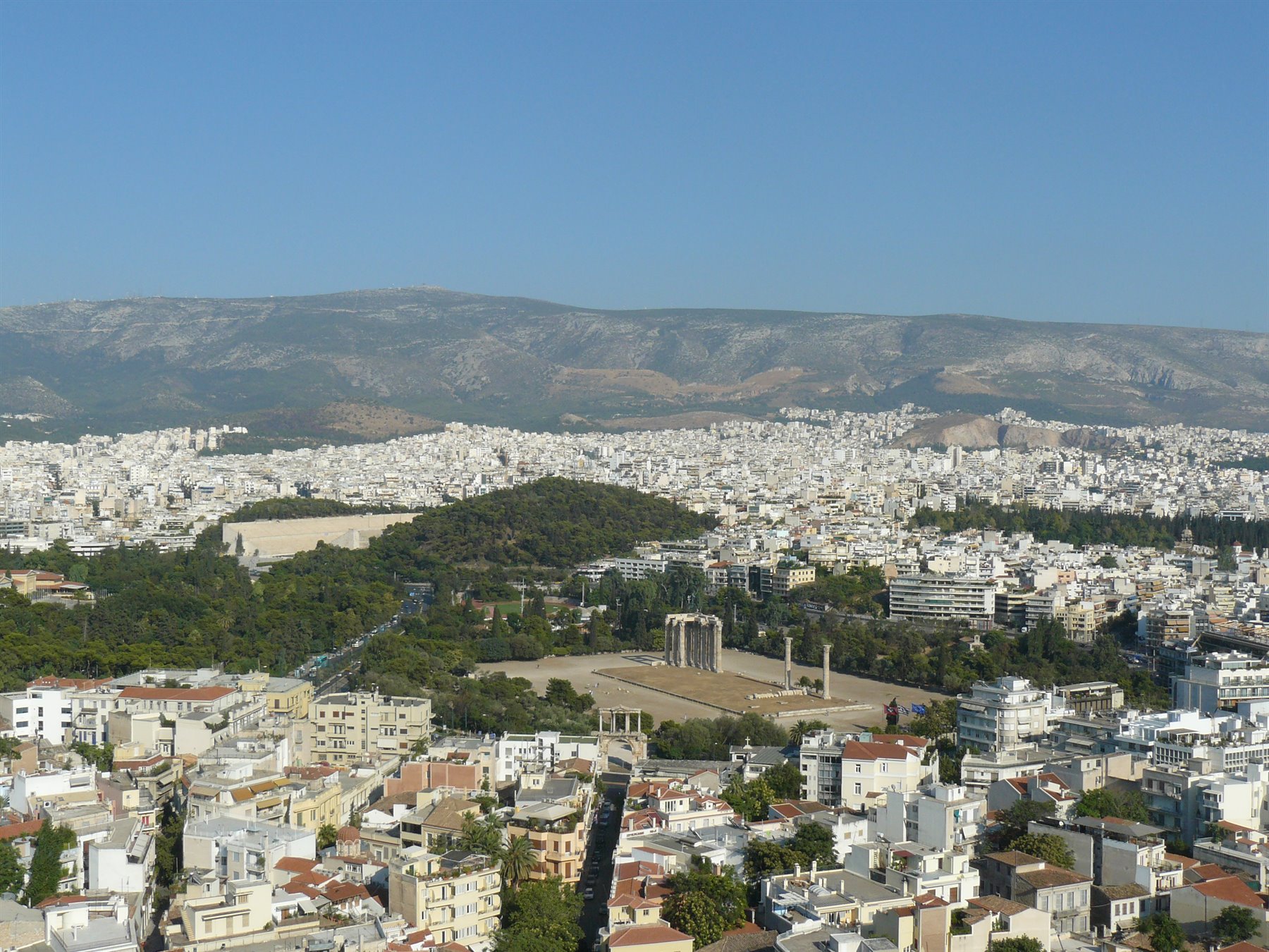 Atene: case bianche, colonne antiche e collina verde sotto monti aridi