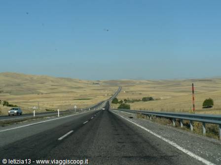 Straight highway through dry yellow hills, metal guardrails, pale blue sky
