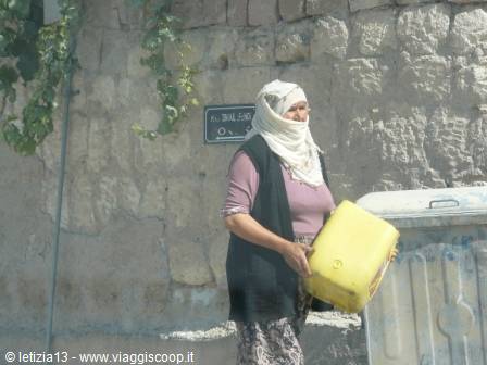Woman in white headscarf carries a yellow jerrycan beside a stone wall