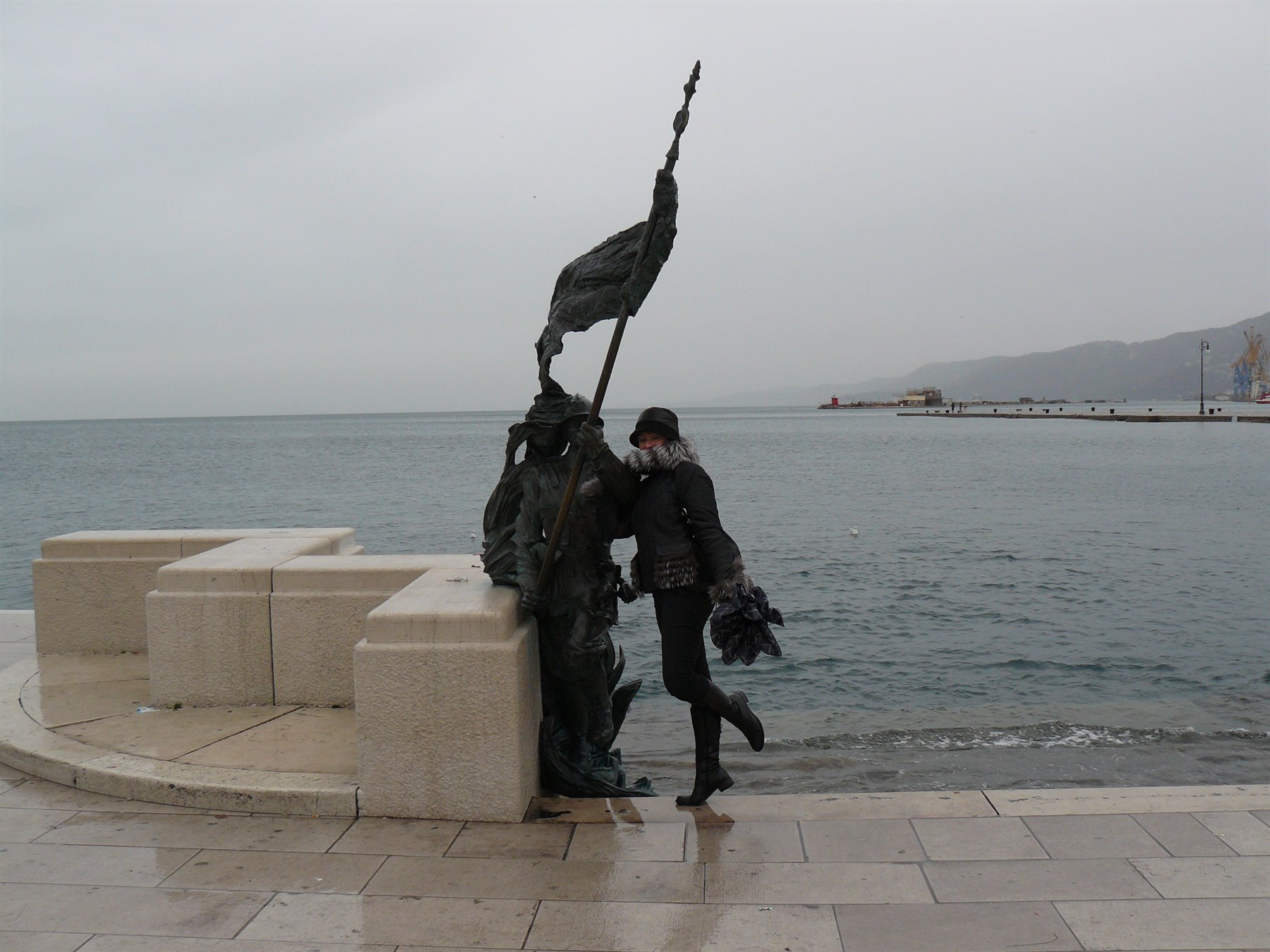 Turista posa accanto a una statua di bronzo sul molo di pietra, cielo grigio