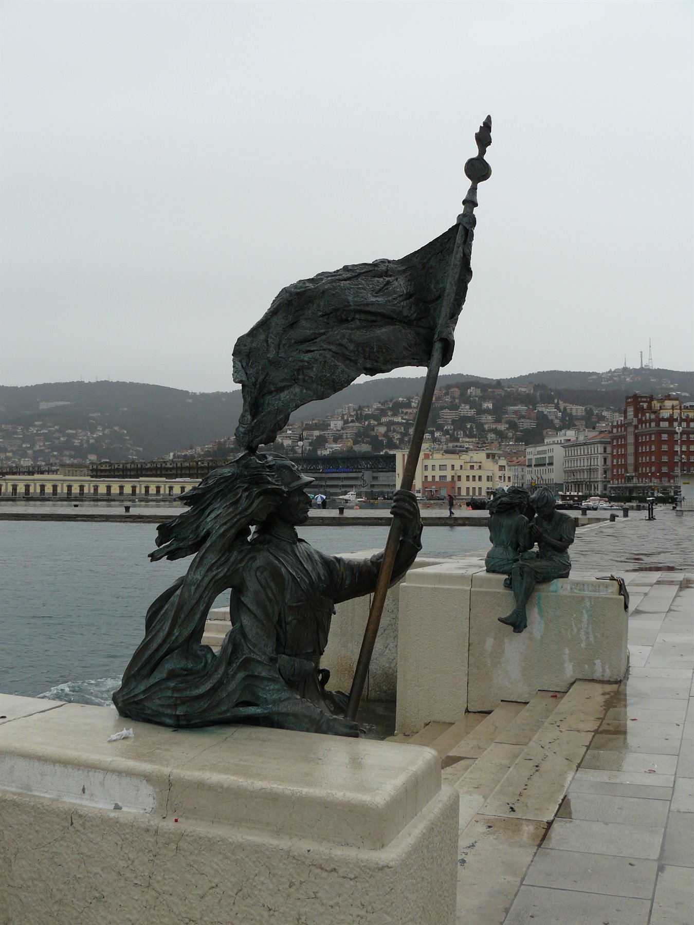 Statua in bronzo con bandiera sul molo di Trieste, cielo grigio
