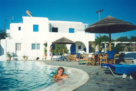 Swimmer in pool beside whitewashed hotel with straw umbrellas and loungers